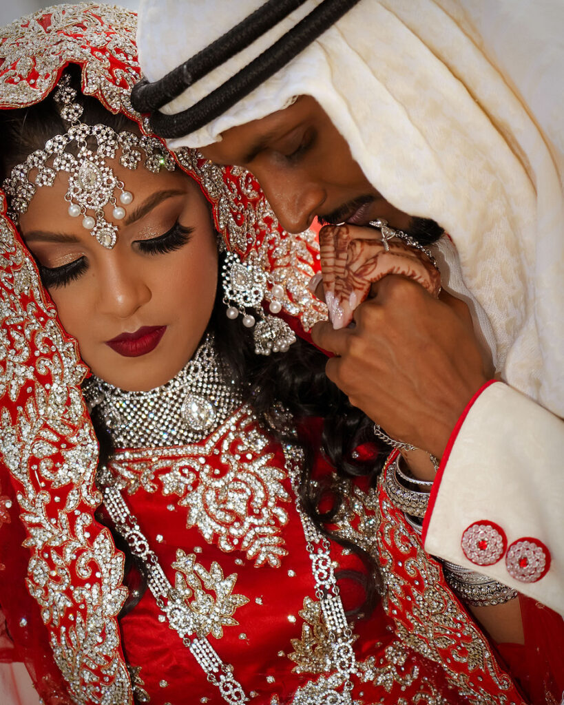 Pakistani groom kissing bride’s hand on wedding day, both in traditional wedding clothing, romantic portrait.