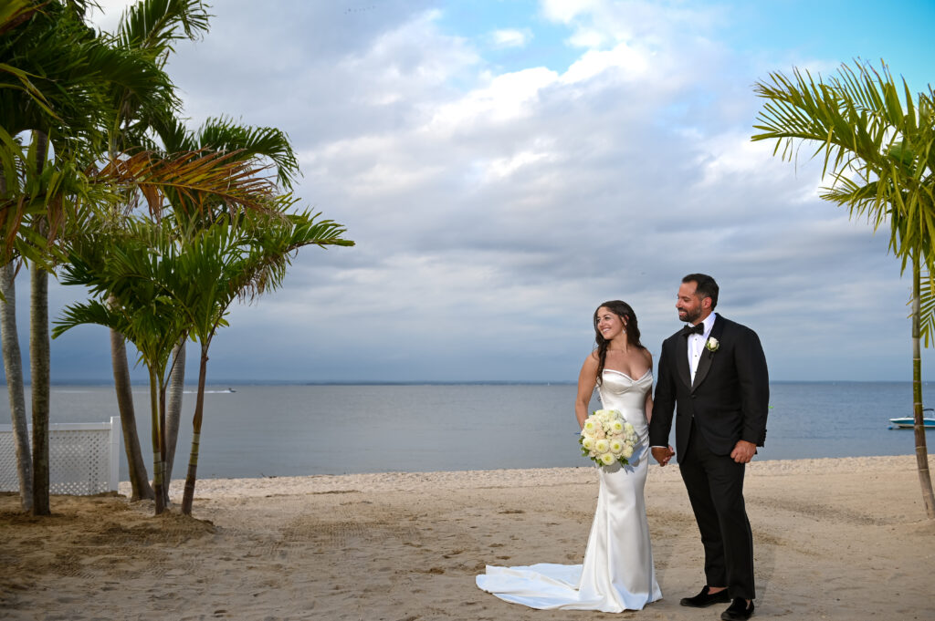 Bride and groom standing on the beach with palm trees at Crescent Beach Club, Long Island, New York.